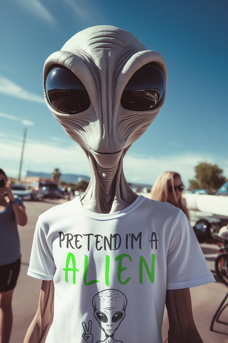 May include: A person wearing an alien mask and a white t-shirt with the text "PRETEND I'M A ALIEN" in green. The shirt also features a cartoon alien illustration. The background includes a blue sky and other people.