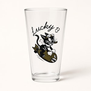 May include: A clear glass pint tumbler featuring a black and olive green graphic of a cartoon cat wearing goggles and riding a bomb. The text "lucky 13" is printed in black script above the cat. The cat has white paws and a white scarf.