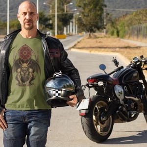 May include: An olive green t-shirt featuring a detailed illustration of a gargoyle with wings and a stained-glass window design. The person is wearing a black leather jacket, blue jeans, and holding a black and white checkered helmet. A black motorcycle is parked nearby.
