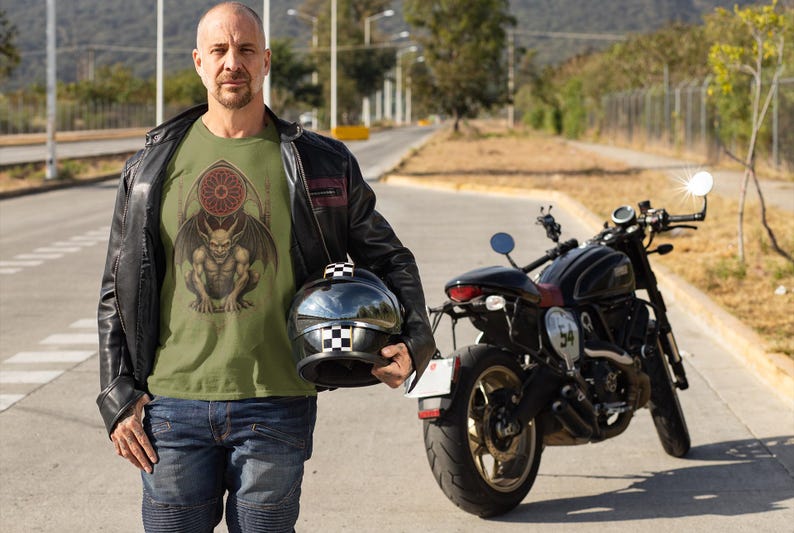 May include: An olive green t-shirt featuring a detailed illustration of a gargoyle with wings and a stained-glass window design. The person is wearing a black leather jacket, blue jeans, and holding a black and white checkered helmet. A black motorcycle is parked nearby.