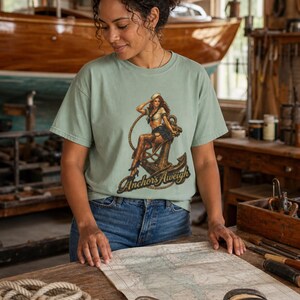 May include: A sage green short-sleeve t-shirt featuring a graphic print of a pin-up sailor sitting on a large metal anchor with a rope. The text "Anchors Aweigh" is printed in a dark, stylized font at the bottom of the graphic design.