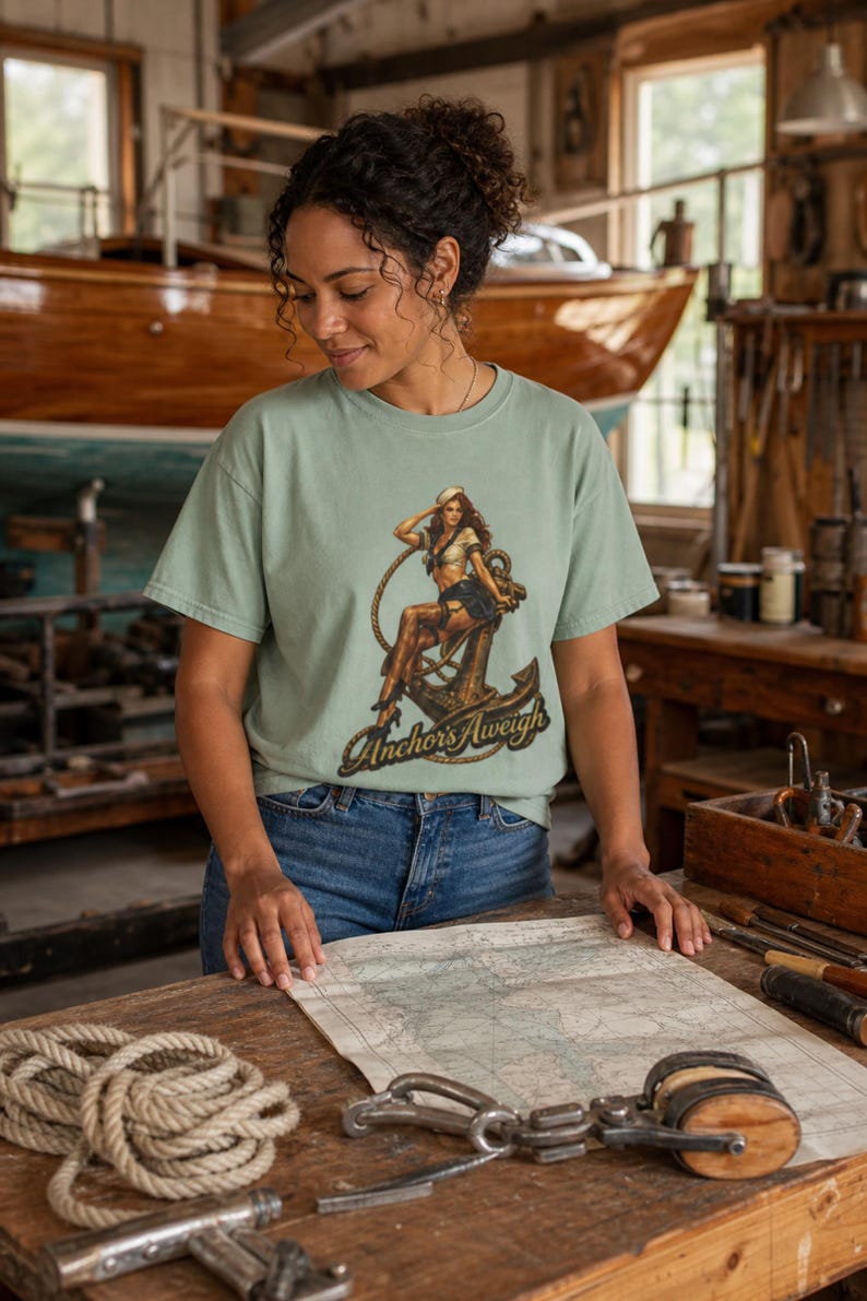 May include: A sage green short-sleeve t-shirt featuring a graphic print of a pin-up sailor sitting on a large metal anchor with a rope. The text "Anchors Aweigh" is printed in a dark, stylized font at the bottom of the graphic design.