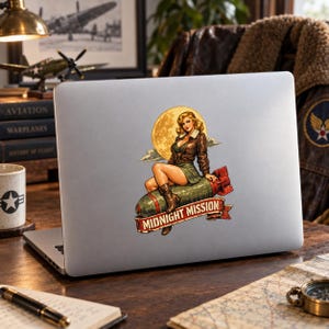 May include: A silver laptop featuring a sticker of a pin-up style illustration. The sticker depicts a person in a brown leather jacket and green skirt sitting on a green bomb against a yellow moon, with a red banner reading MIDNIGHT MISSION.
