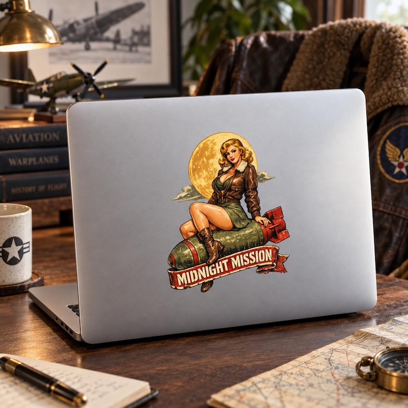 K&ouml;nnte beinhalten: Ein silberner Laptop mit einem Aufkleber im Pin-up-Stil. Der Aufkleber zeigt eine Person in einer braunen Lederjacke und einem gr&uuml;nen Rock, die auf einer gr&uuml;nen Bombe vor einem gelben Mond sitzt, mit einem roten Banner mit der Aufschrift MIDNIGHT MISSION.