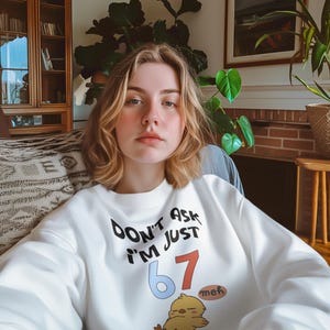May include: White sweatshirt with the text "Don't Ask I'm Just 67 meh" in black, red, and blue. A cartoon chick is printed below the text. The sweatshirt has long sleeves.