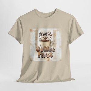 May include: A beige short-sleeve t-shirt featuring a central graphic of a coffee mug with steam and the text "Pause Coffee First" in a brown, layered font. The background of the graphic consists of vertical stripes in varying shades of tan and white.