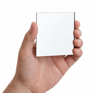 May include: A square mirror with a black edge is held in a hand against a white backdrop. The mirror's surface is reflective and clean. The image is a close-up shot, highlighting the mirror's reflective qualities.