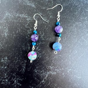 May include: A pair of dangle earrings with silver hooks. Each earring has round beads in purple, blue, and patterned pink and blue. The beads are strung together with silver wire. The earrings are suitable for everyday wear.