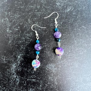 May include: Dangle earrings with a silver hook, featuring a combination of purple, blue, and white beads. The earrings are made up of round beads in different sizes and colours, creating a layered effect. The beads have a marbled appearance.