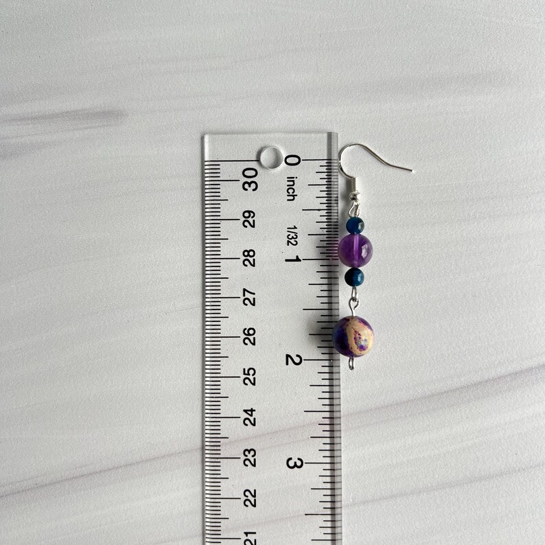 May include: A single earring with a silver hook, featuring a series of beads. The earring includes a dark purple, a light purple, and two blue beads. The bottom bead is a cream colour with purple and brown swirls. The earring is approximately 5 cm long.