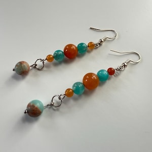 May include: A pair of dangle earrings featuring a series of colorful beads. The earrings have a silver-tone hook and are adorned with a variety of beads in shades of orange, turquoise, and brown. The beads vary in size and shape, creating a unique and eye-catching design.