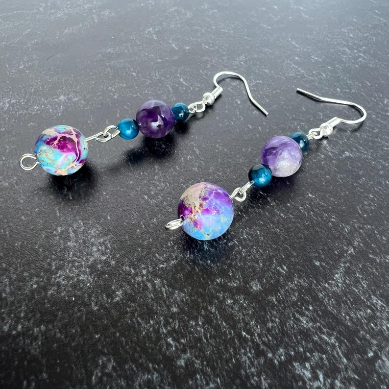 May include: Dangle earrings with a mix of colourful beads. Each earring has a large, round, speckled bead in purple, blue, and pink. The earrings also include a purple amethyst bead and smaller blue beads, all connected with silver-tone metal.