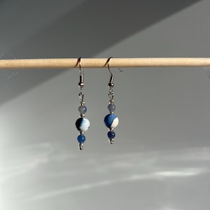 May include: A pair of dangle earrings with silver-colored hooks. Each earring features a small, light blue bead, followed by a larger, round bead with a white and blue swirl pattern, and a smaller, dark blue bead. The earrings are displayed on a wooden dowel.