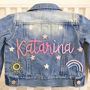 May include: A blue denim jacket with the name "Katarina" embroidered in pink script on the back. The jacket is decorated with embroidered flowers, a sun, and a rainbow. The jacket has button closures and a collar.