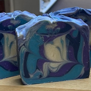 May include: Three handmade soap bars with a marbled design. The soap features swirls of blue, purple, and cream colors. The bars are rectangular and have a textured, uneven top surface. The soap is displayed on a wooden surface.