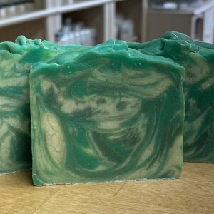 May include: Three handmade soap bars with a green and white swirl pattern. The soap has a textured surface and a natural, organic appearance. The bars are displayed on a wooden surface.