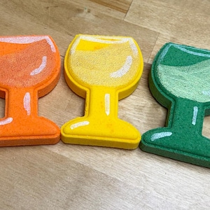 May include: Five brightly coloured wine glass-shaped objects in pink, orange, yellow, green, and blue. Each has a white outline and a textured appearance, possibly sponge-like. The glasses are arranged diagonally on a light brown surface.