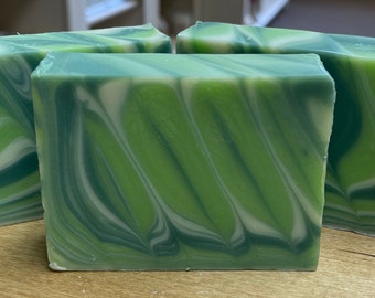 May include: Three rectangular bars of handmade soap with a green, white, and dark green swirled pattern. The soap bars are arranged on a light brown surface, showcasing the marbled design. The soap appears to be freshly cut.