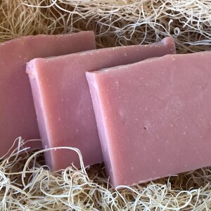 May include: Three rectangular bars of pink soap are arranged on a bed of dried, light-brown plant fibers. The soap has a smooth surface and a slightly uneven edge, suggesting a handmade quality. The overall presentation is natural and rustic.