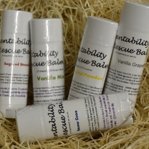 Puede incluir: Cinco tubos blancos de bálsamo labial con etiquetas que dicen "Rescue Balm" y varios sabores como "Sugared Strawberry" y "Vanilla Grape". Los tubos están dispuestos sobre un lecho de material similar a la paja.