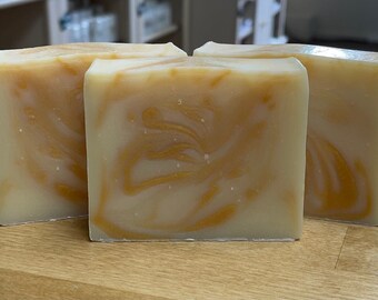 May include: Three rectangular bars of handmade soap. The soap has a creamy white base with golden-yellow swirls throughout. The bars are sitting on a light brown wooden surface.