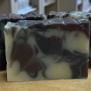 May include: Four rectangular bars of handmade soap with a marbled design. The soap features a combination of white, brown, and dark brown swirls. The bars are arranged on a wooden surface.
