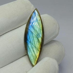 Peut inclure: Une pierre précieuse polie et allongée en forme de goutte d'eau. La pierre présente un jeu de couleurs vibrant, avec des teintes de bleu, de vert et de jaune. La surface présente un motif subtil et texturé. Le bijou est tenu par une main gantée.