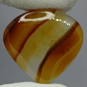 Puede incluir: Una piedra preciosa de ágata pulida en forma de corazón con bandas translúcidas de color ámbar, amarillo dorado y marrón rojizo intenso. La piedra tiene una superficie lisa y brillante con patrones naturales y un reflejo blanco distintivo.