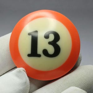 Puede incluir: Una bola de billar con el número 13 en negro, centrado en un círculo crema. El círculo está rodeado por una banda naranja brillante. La bola es de tamaño estándar para juegos de billar.