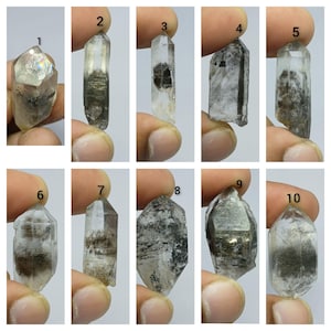 Könnte beinhalten: Eine Sammlung von zehn klaren Quarzkristallen, nummeriert von eins bis zehn, mit ausgeprägten dunklen Einschlüssen, unterschiedlichen geometrischen Formen und spitzen Enden, einzeln vor einem schlichten weißen Hintergrund dargestellt.