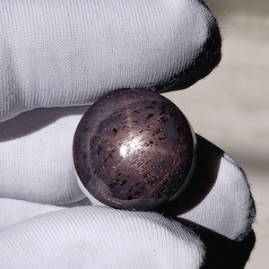 May include: A polished, dark purple gemstone sphere with a speckled pattern. The sphere is held in a white gloved hand, showcasing its smooth, reflective surface. The gemstone appears to be approximately 3.8 cm in diameter.