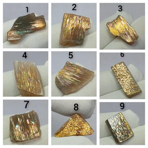 May include: A collection of nine numbered sunstone specimens, each displaying unique internal golden, orange, and iridescent metallic inclusions. These raw, polished-face gemstones exhibit varying shapes and light-reflecting patterns across their surfaces.