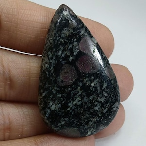 May include: A teardrop-shaped gemstone with a dark, speckled background and several deep red inclusions. The polished stone has a smooth, glossy surface, and the inclusions are irregularly shaped. The overall color is a mix of black, gray, and red.