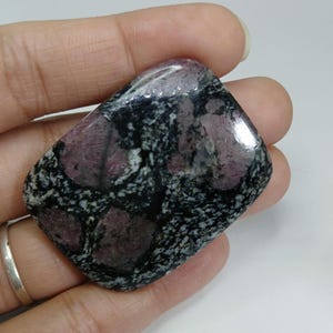 May include: A polished gemstone with a mix of colors. The stone features a dark, speckled background with patches of deep red and a glossy finish. The shape is rectangular with rounded edges.