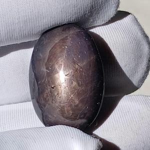 May include: A dark, purplish-brown oval gemstone with a polished surface. Light reflects off the surface, creating a star-like pattern. The gem is held by a gloved hand, emphasizing its smooth, reflective quality. A beautiful example of natural mineral beauty.