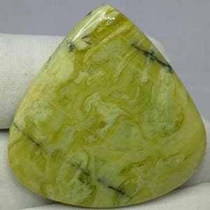Puede incluir: Una piedra preciosa pulida de forma triangular con un patrón jaspeado en tonos chartreuse y amarillo pálido, con finas inclusiones oscuras similares a vetas en la superficie lisa y brillante de la piedra.