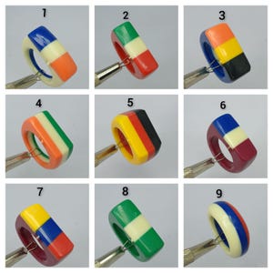 May include: A collection of nine chunky resin rings, each featuring a distinct three-color block pattern. The rings are numbered one through nine and are displayed individually, held by a metal tool to showcase their smooth, glossy finish.