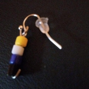 May include: A single dangle earring featuring a silver-toned metal hook with a clear plastic backing. The earring is decorated with a vertical stack of four small cylindrical beads in yellow, white, purple, and black colors arranged in a sequence.