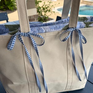 May include: A beige canvas tote bag featuring two sturdy shoulder straps and a blue and white gingham ribbon tied in bows at the top. The interior lining displays a floral pattern in shades of blue and green, visible along the upper edge.