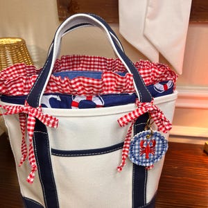 May include: A white canvas tote bag with navy blue accents and handles. The bag features a red and white gingham lining and trim. A blue and white gingham lobster charm is attached with a red and white gingham ribbon. The bag has a nautical theme.