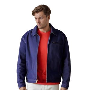 Può includere: Giacca blu navy con zip e colletto, indossata sopra una t-shirt rossa. La giacca ha una piccola tasca sul petto e due tasche laterali. Il modello indossa pantaloni bianchi. La giacca è realizzata in materiale resistente.