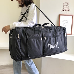 May include: A black travel duffel bag with multiple zippered compartments and a shoulder strap. The word "TRAVEL" is printed in white on the front. The bag is being carried by a person.