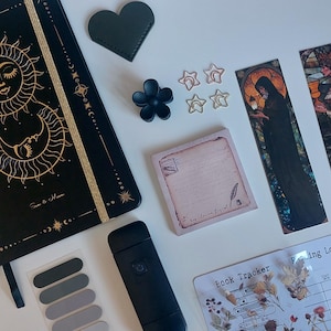 Op de afbeelding: Een zwart dagboek met gouden zon- en maanillustraties, twee rechthoekige bladwijzers met glas-in-loodstijl kunst, een vierkant notitieblok met vintage briefmotieven, een zwarte haarklem, vier stervormige paperclips en een boektrackerblad.