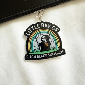 May include: A black and white keychain with the text "LITTLE RAY OF PITCH BLACK SUNSHINE". The keychain features a cartoon Grim Reaper character with a scythe under a rainbow.