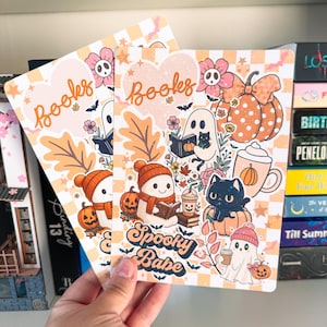 May include: Two decorative cards with a fall theme. The cards feature illustrations of ghosts, pumpkins, and cats, with the word "Books" and "Spooky Babe" on them. The background is a checkered pattern in orange and white.
