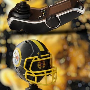 May include: Black and yellow Pittsburgh Steelers football helmet miniature on a black joystick controller. The helmet has the Steelers logo on the side and a yellow facemask. The controller has a screen and buttons.
