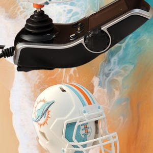 May include: Two miniature white football helmets with the Miami Dolphins logo, featuring aqua and orange stripes. The helmets are mounted on a black joystick and controller, set against a colorful abstract background.
