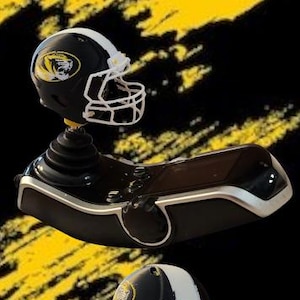 May include: A black and silver gaming joystick controller featuring a miniature black football helmet with a white face mask and a yellow tiger logo on the side, mounted on the top of the joystick handle, shown from two different angles.