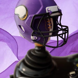 May include: A purple and white miniature football helmet with a black facemask. The helmet is mounted on a black base, likely a joystick. The helmet features the logo of the Minnesota Vikings.