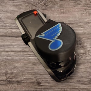 May include: A black and silver electronic device with a round black top featuring a blue and light blue logo. The device has a screen and an orange accent. The item is on a wooden surface.